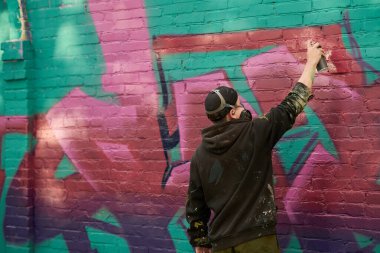 Genç bir adamın yeşil tuğla duvara mor boya sıkarken sokakta yeni graffiti çizimleri yaptığı arka plan.
