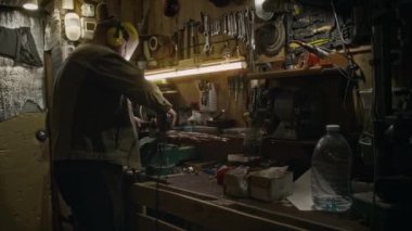 Orta boy koruyucu üniformalı erkek teknisyen, elektrikli testere tutan kulaklıklar, atölyeye sabitlenmiş metal parçaları, uçan kıvılcımlarla kesip parlatıyorlar.