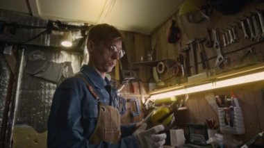 Kafkasyalı genç tamircinin önlüklü orta boy fotoğrafı, eldivenler kulaklık takıyor, koruyucu şapka takıyor ve loş ışıklandırılmış garajda veya atölyede metalle çalışıyor.