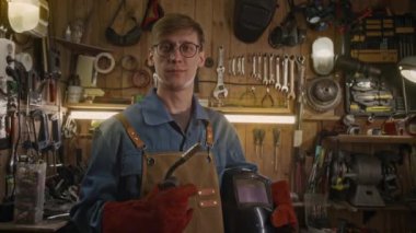 Orta ölçekli mutlu, beyaz erkek kaynakçı koruyucu deri eldivenler, kask ve kaynak makinesi, atölye veya garajda duvarın önünde duran araçlar