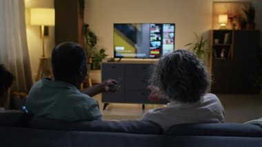 Kanepede çocukla oturan ve TV 'de hangi filmi izleyeceklerini tartışan, uzaktan kumandayı birbirlerinden alan, evdeki yayın servisini kullanan evli çiftlerin arka görüntüsü.