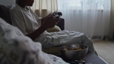 Evdeki oyun bahçesinde hevesle video oyunu oynarken kanepeye oturmuş patates cipsi yiyen anonim, koyu tenli bir çocuğun görüntüsü.