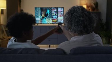 Farklı ailelerin TV 'de birlikte film izlemesi, çocuğun annesinden uzaktan kumanda alması ve yayın servisinde ne izleyeceğini seçmesi, modern oturma odasındaki kanepede oturması.