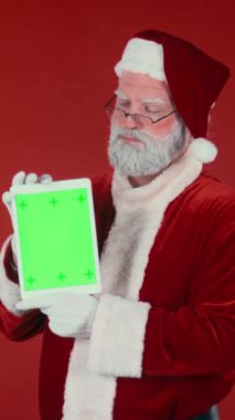 Geleneksel kostümlü karizmatik Noel Baba 'nın dikey orta saplama portresi dijital tablet taşıyor ve krom anahtar yeşil ekrana bakıyor, kırmızı arka planda kameraya bakıyor, boşluğu kopyalıyor