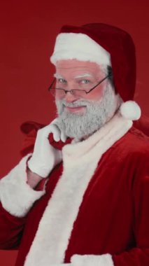 Geleneksel kostümlü mutlu Noel Baba 'nın dikey göğsünden bıçaklanmış portresi ve alarm saatini gösteren çuval, kırmızı arka planda duran çocuklara hediyeler dağıtmaya hazır.