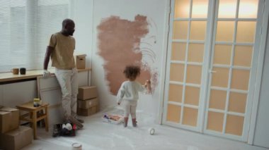 Afrikalı Amerikalı bir babanın dinlenme ve melez kızının ona yardım edişini izlediği, yeni dairesinin bakım odasının yenilenmesi sırasında duvarları mini silindirle boyadığı bir fotoğraf.