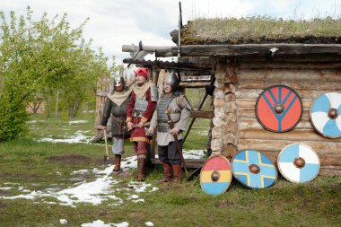 Ortaçağ savaşçı kostümlü bir grup tarihi canlandırıcı ahşap bir kulübenin önünde duruyor ve kalkanları var. Yeşil çevre ile zemin kontrastında seyrek kar yamaları