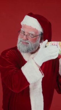 Geleneksel kostüm sallayan modern Noel Baba 'nın dikey orta boy portresi stüdyoda kırmızı arka planda poz verirken onu size sunuyor. Şablon, boşluğu kopyala
