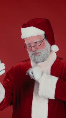 Noel Baba 'nın gözlüklü ve geleneksel kostümlü, modern akıllı telefondan gülen ve kırmızı arka planda konuşan modaya uygun fotoğrafının dikey orta kesimi. Şablon için uygun, alanı kopyala