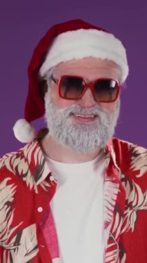 Dik Göğüs hizasında, güneş gözlüklü, kırmızı şapkalı ve aloha gömlekli rahat Avustralyalı Noel Baba 'nın portresi. İki eli de mor arka planda serçe parmak işareti yapıyor ve kameraya bakıyor.