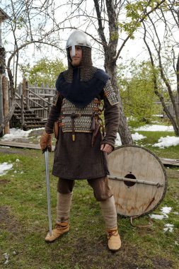 Medieval knight standing on grass while holding sword in hand, armored and ready in traditional garb, with wooden shield in background and trees framing scene clipart