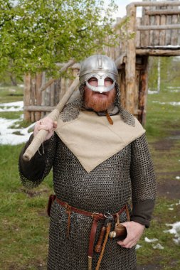 Viking canlandırmasının portresi geleneksel zincir ve miğfer giyerken açık hava tarihi ortamında tahta silah tutuyordu. Arkaplanda ağaçlar ve sakallı, gülümseyen bir adam.
