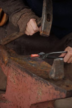 Geleneksel demircilik aletleriyle çalışan yetenekli bir zanaatkar örs üzerinde metal ısıtıyor. Yakın çekim, el ve ekipman gösterimi, detaylar ve işçiliğin vurgulanması