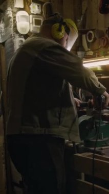 Korumalı takım elbiseli erkek teknisyenin orta boy dikey görüntüsü. Kulaklıklar elektrikli testereyle metal parçalarını parlatırken garajda çalışıyor.