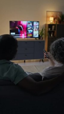 Kanepede kucaklaşan ve akşam oturma odasında TV 'de hangi filmi izleyeceklerini seçen çeşitli eşlerin dikey arka çekimi.