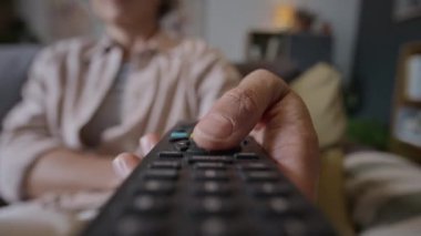 Uzaktan kumandayı elinde tutan, düğmelere basan, evde televizyon seyrederken kanal değiştiren tanınmamış bir kadının yakın çekimi.