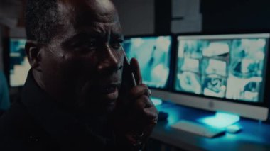 Kaş çatan, endişeli, ev telefonuyla iş arkadaşı veya müdürüyle konuşan siyah güvenlik görevlisinin kapanışı, CCTV görüntülerini incelerken olası bir olayı rapor ediyor.