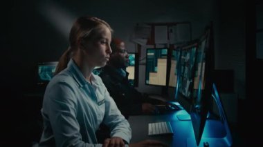 Kontrol odasında çalışan iki etnik çeşitliliğe sahip güvenlik görevlisinin orta ölçekli görüntüsü, beyaz kadın şüpheli hareketler fark ediyor, CCTV ekranını işaret ediyor, telsizle devriyeyi arayan siyahi adama bildiriyor.