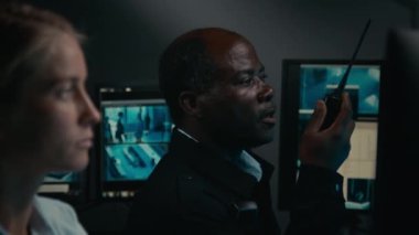 Orta yaşlı Afro-Amerikan güvenlik ajanının yakın plan çekimleri portatif telsizle ya da yer devriyesiyle konuşurken, şüpheli hareketler bildirirken ve kadın meslektaşı CCTV izlerken.