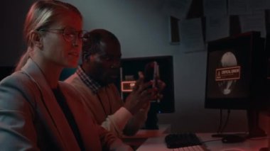 Siyahi erkek ve beyaz kadın bilişim uzmanlarının şirket sistemine saldırıyı savuşturmak için savaştığı, telefonla tehdit bildirdiği, tartıştığı ve ekranlara işaret ettiği orta ölçekli bir fotoğraf.