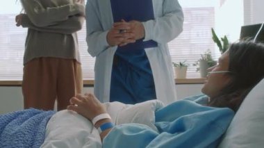 Burun kanülü beyaz bir kızın, gözlüklü bir erkek doktorla konuşurken, annesinin ayakta durup hastane koğuşunda dinlediğini gösteren bir fotoğraf.