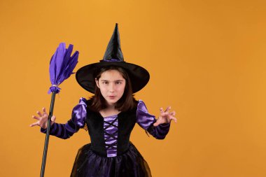 Siyah şapkalı ve elbiseli şirin küçük cadı mor süpürge tutuyor ve turuncu arka planda hırıldarken kameraya bakıyor.