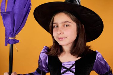 Süslü kostümlü gülümseyen genç cadı elinde mor süpürgeyle turuncu arka planda kameraya bakıyor.