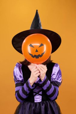 Siyah şapkalı küçük bir cadının kamera önünde turuncu arka planda poz verirken yüzünü balkabağı balonuyla kaplaması.