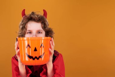 Kırmızı boynuzları olan, turuncu balkabağı şeklindeki plastik kovayı dikizleyen tatlı bir çocuk.