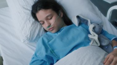 Hastane yatağında uzanmış, hasta yatağında uzanmış, klinikte yanında otururken ailesinin elini tutmasından endişe duyan baygın bir kız.