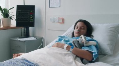 Küçük beyaz kızın hastane yatağında tavşan pelüş, burun uzaması ve klinikteki iyileşme sürecinde kolunda serumla uyuduğu bir resim.