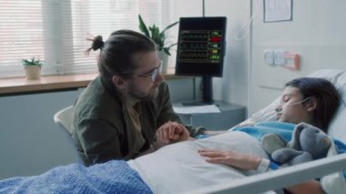 Gözlüklü, orta karar beyaz bir adam hastane yatağında, çocuk hastanesinde baygın bir kızın elini tutuyor.