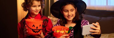 Panorama of happy girl in Halloween costumes sitting on bed, holding buckets with candies and taking selfie on smartphone clipart