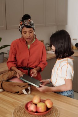 Two women standing by wooden counter engaging in conversation while holding books. Fruit bowl with apples adding touch of home in warm kitchen space clipart