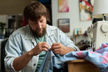 Sakallı erkek işçi ya da ikinci el kot pantolon satan ikinci el dükkan sahibi iş yerinde dikiş makinesiyle otururken