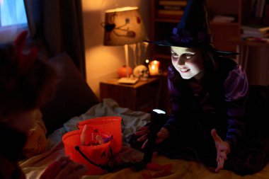 Cute little girl in Halloween witch costume shining flashlight on her face while sitting on bed in front of boy and telling him horror story clipart