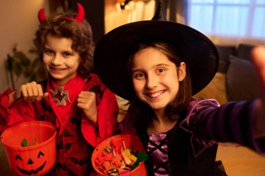 Siyah şapkalı ve cadı elbiseli tatlı bir kız. Kardeşinin yanında oturuyor. Akıllı telefondan fotoğraf çektirirken şeytan kıyafeti giyiyor.