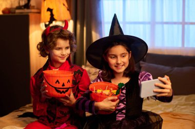 Happy girl in witch dress and boy in devil costume sitting on bed, holding buckets with candies and taking selfie on smartphone clipart