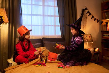 Side view of two friendly kids in Halloween outfit sitting on bed against large bedroom window and sharing candies clipart