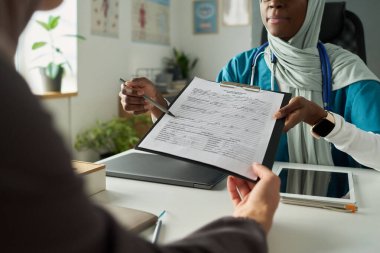 Ofis ortamında iki Müslüman kadın, biri bir diğerine danışmanlık yaparken not alıyor. Kağıtlar, cihazlar ve bitkiler profesyonel ve sakin bir ortam oluşturur