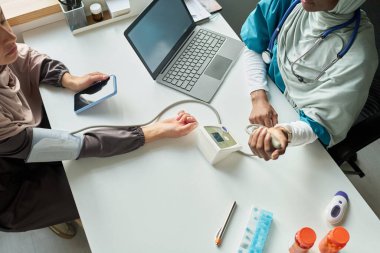 Muslim medical professional measuring patient blood pressure with digital device in healthcare setting clipart