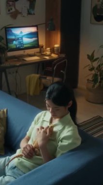 Evdeki kanepede uzanmış yeşil polo tişörtlü, göğsünde sakallı agama kertenkelesi tutan, başını okşayan düşünceli, yalnız Çinli kadının orta açılı yüksek açılı görüntüsü.