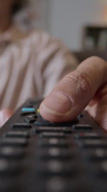 Uzaktan kumandalı, düğmelere basan, evde televizyon seyrederken kanal değiştiren, tanınmayan bir kadının dikey yakın çekimi.