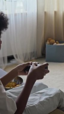 Küçük siyahi çocuğun evdeki plazma TV 'de atış oyunu oynarken kanepede oturduğu dikey yay çekimi.