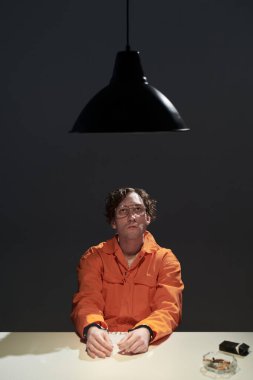 Portrait of prisoner wearing orange jumpsuit sitting alone under hanging lamp, focusing on folded hands placed on table. Ashtray with cigarette butts visible across table clipart