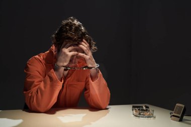 Turuncu tulumlu şahıs elleri bağlı masada oturuyor, kafası derin düşüncelere dalmış ya da endişeli gibi elleri üzerinde dinleniyor. Görünür kelepçeler yerçekimi senaryolarını vurguluyor