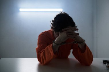 Turuncu tulum giyen bir adam elinde kafasıyla masada oturuyor, floresan ışıkla aydınlatılıyor, dramatik gölgeler ve yansıma atmosferi yaratıyor.