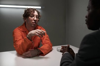 Turuncu tulumlu bir mahkum, elleri kelepçeli, sorgu sırasında loş ışıklı bir odada Afrikalı Amerikalı memurla sohbet ediyor.