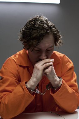 Düşünceli adam turuncu tulum ve kelepçe takıyor masada tek başına oturuyor, elleri üzerinde dinleniyor, düşünceli ve parlak ışıklı bir odada odaklanmış görünüyor.