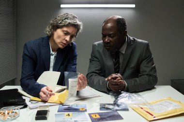 Delilleri dikkatle inceleyen iki dedektif, soruşturma sırasında evrak ve aletlerle dolu loş bir odada bulguları tartışıyor. İkisi de odaklanmış ve ciddi görünüyor.